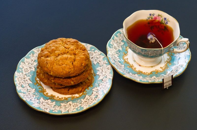 A stack of Anzac Biscuits on a china plate next to a cup of black English Breakfast tea