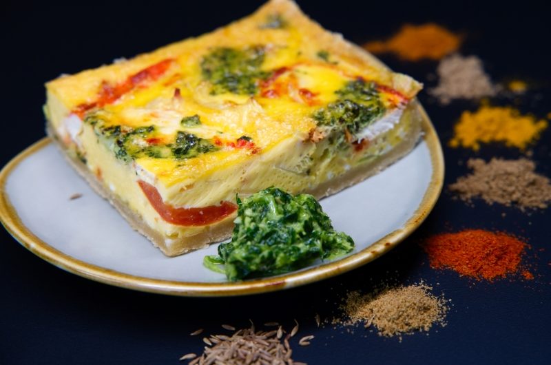 A square slice of chicken tikka flavoured quiche on a plate surrounded by 7 piles of the different spices used to flavour the chicken