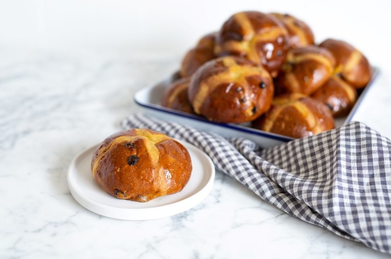 Hot Cross Buns 2021 A round hot cross bun on a plate with a stack of hot cross buns in the background
