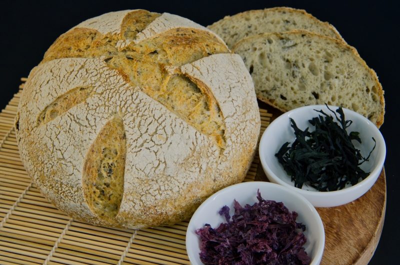 Round loaf of sourdough bread with two slices cut and two small bowls of seaweed flakes and dried seaweed